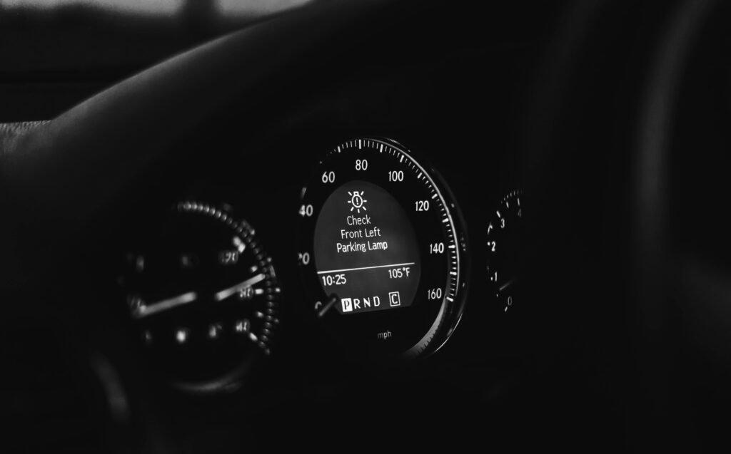 Close-up of a car dashboard in black and white, highlighting speedometer and warning lights.